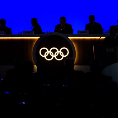 Olympiska ringarna lyser i ett bord. Vid bordet ser man en mörk siluett av internationella olympiska kommittén.