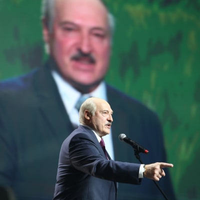 Aleksandr Lukasjenko står i en talarstol och pekar framåt med ett finger. I bakgrunden ett stort fotografi på honom. Den 17 september 2020.