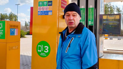Man i blå jacka står framför orange bensinpump.