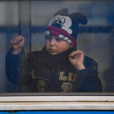 En pojke i mussepiggmössa tittar ut genom ett smutsigt tågfönster.