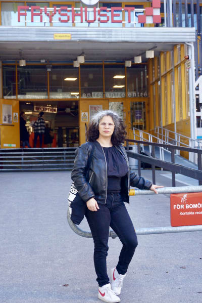 En kvinna med svart och grått lockigt hår står utanför en byggnad med texten Fryshuset