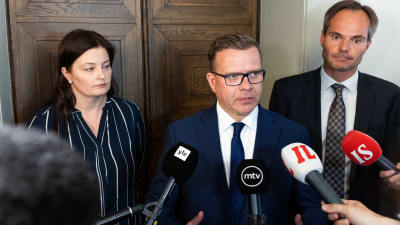 Samlingspartiets partisekreterare Kristiina Kokko, ordförande Petteri Orpo och riksdagsgruppens ordförande Kai Mykkänen står framför en dörr. I förgrunden händer med framsträckta mikrofoner.