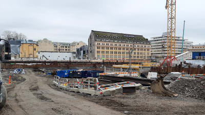 Byggkonstruktioner, ställningar, lastbilsflak och en byggkran står i gropen till det som byggs till en torgparkering under Salutorget i Åbo.