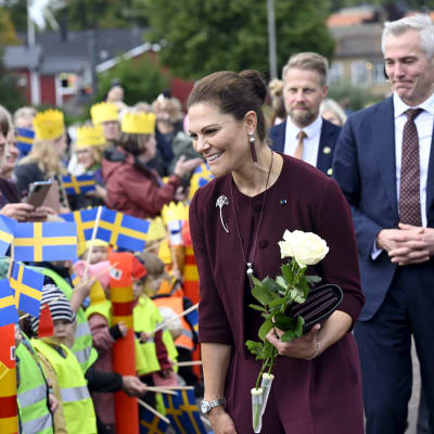 Kronprinsessan Victoria ler och hälsar på en grupp barn som viftar med svenska flaggor. Flera av barnen har gula papperskronor på huvudet. I sin hand har kronprinsessan ett par vita rosor och bakom henne går Anders Adlercreutz.
