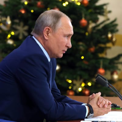 Vladimir Putin sitter vid sitt bord i sidoprofil och har en dekorerad julgran i bakgrunde. Putin har knäppt sina händer.