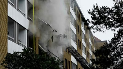 Rök stiger från ett flervåningshus, brandbilar bredvid.
