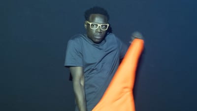 En man i glasögon står framför en blå vägg. Han tittar åt sidan och viftar med en orange flagga.