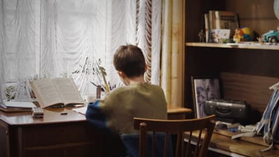 En pojke fotograferad bakifrån medan han sitter och jobbar vid ett skrivbord och läser.