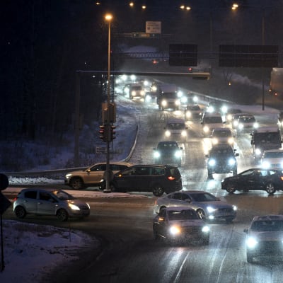 Mycket bilar i en korsning vid en stor väg i snöigt och mörkt väder.