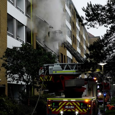 Rök stiger från ett flervåningshus, brandbilar bredvid.