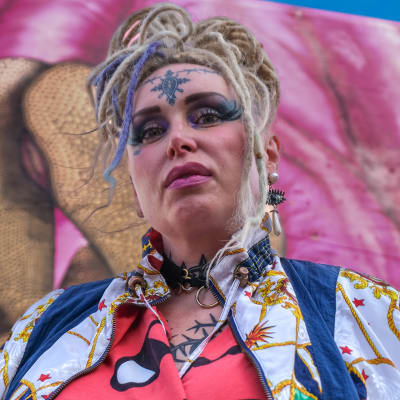 Kvinna i färggranna kläder, blond rastafrisyr och tatueringar i ansiktet tittar in i kameran. I bakgrunden en stor mural som föreställer kvinnligt könsorgan.