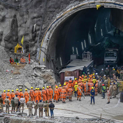  Ett fyrtiotal arbetare i orange overaller står och köar vid mynningen till en tunnuel. Intill tunneln står en grupp på ett drygt tiotal personer i camouflageoveraller.