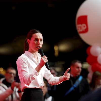Sanna Marin med allvarlig min håller tal på valkampanjtillfälle, med SDP-ballonger i bakgrunden