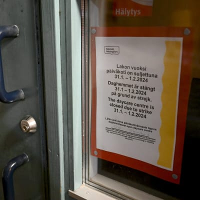 En lapp där det står "Daghemmet är stängt 31.1 – 1.2.2024 på grund av strejk" uppsatt i ett fönster bredvid en grå dörr.