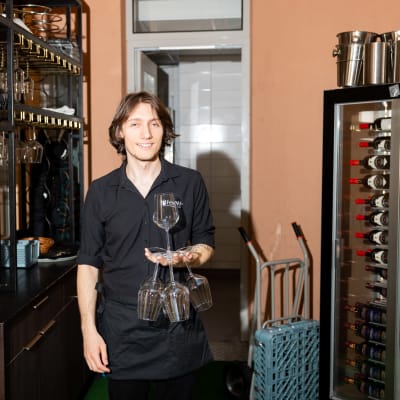 Tedo Kartsivadze pitää kädessään kuutta viinilasia. Taustalla näkyy keittiön ovi ja oikealla puolella on viinikaappi.