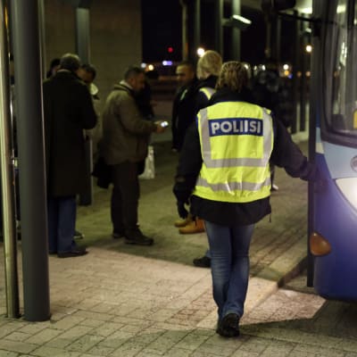 En polis med neongul väst står vid en buss och väntar på att en grupp asylsökande ska stiga på.