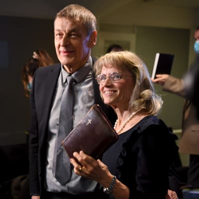 Päivi Räsänen och hennes man ler mot en grupp fotografer. Räsänen håller en bibel i handen.