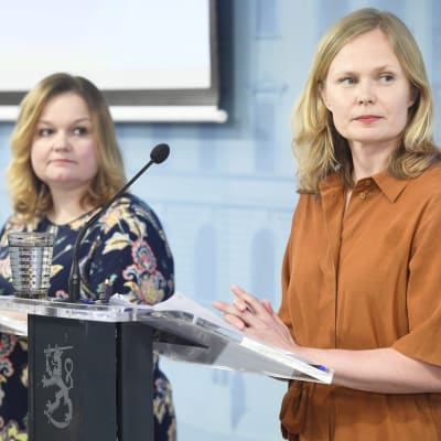 Krista Kiuru och Hanna Kosonen står vid talarpodium framför en blå vägg. De tittar åt sidan.
