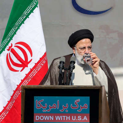 En man i svart turban dricker ett glas vatten. Bredvid honom står Irans flagga och slagordet "Down with USA".