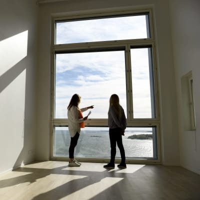 Kaksi henkilöä seisoo seinän kokoisen ikkunan edessä tyhjässä asunnossa. Ikkunasta näkyy meri.