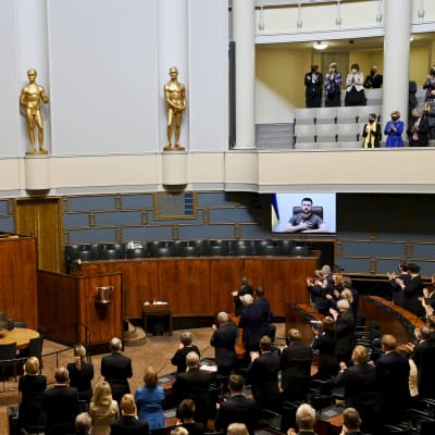 Bild från riksdagen då president Zelenskyj på distans håller tal, via en skärm. Riksdagen står och apploredar.