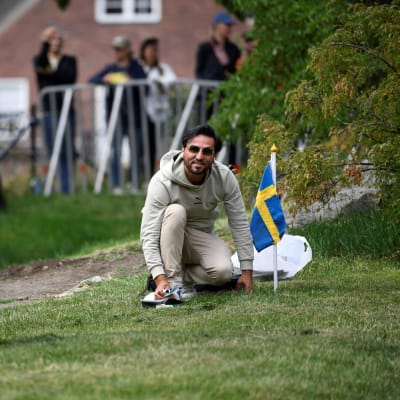 Salwan Momika istuu toinen polvi maassa nurmikolla Ruotsin lippu vieressään ja katsoo kameraan. Vieressä ihminen Magafonin kanssa. Taustalla ihmisiä ja poliiseja.