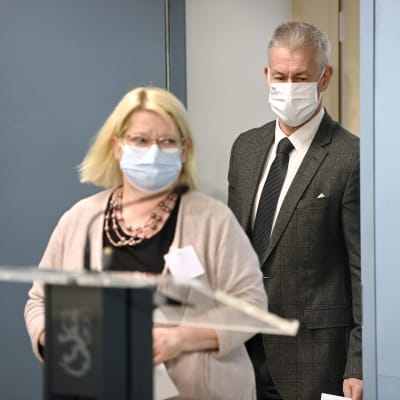 En man och en kvinna i munskydd bakom ett podium på statsrådets presskonferens.