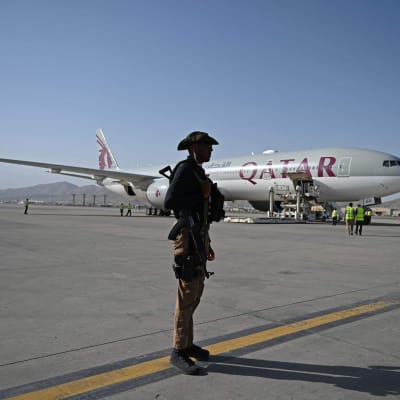 En soldat står på ett flygfält med ett passagerarplan bakom sig. På planet står med stora bokstäver Qatar.
