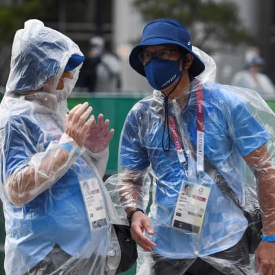 Personal vid OS i Tokyo pratar utomhus. De har på sig regnrockar och munskydd.