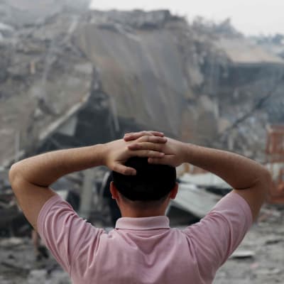 Palestinsk man ser på ett sönderbombat hus och håller händerna bakom huvudet.