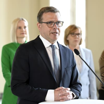Petteri Orpo står vid en mikrofon på en presskonferens, Riikka Purra och Anna-Maja Henriksson står bakom.