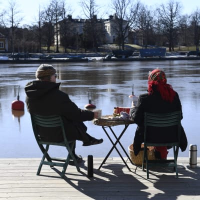 Två personer sitter på stolar med ett litet bord med mat emellan sig. Personerna blickar mot Kajsaniemi i Helsingfors.