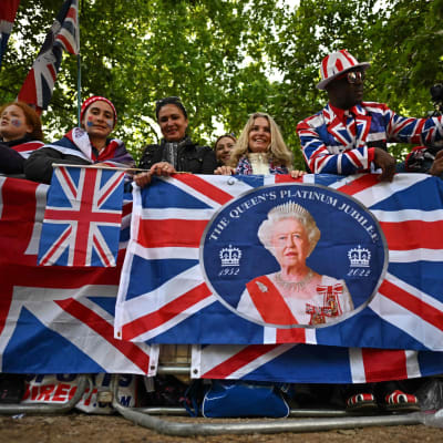 En rad människor med brittiska flaggor och ett porträtt av drottning Elizabeth framför sig. I bakgrunden lövträd.