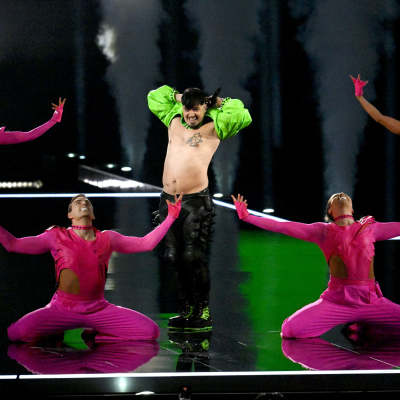 Käärijä seisoo kädet pään takana lavalla vihreässä polerossa ja mustissa housuissa neljän pinkkiin pukeutuneen tanssijan ympäröimänä.