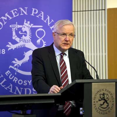 En gråhårig man i kostym talar vid ett podium. Bakom honom en blå plansch med texten Finlands bank och ett lejon. 