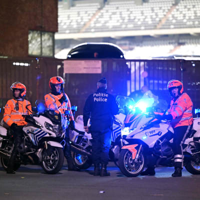 Tre poliser i orangefärgade jackor sitter på var sin motorcykel. En polis i blå uniform står med dem.