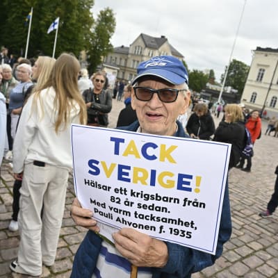 En äldre man i blå keps står på torget i Lovisa och håller i en skylt med texten "Tack Sverige! Hälsar ett krigsbarn från 82 år sedan. Med varm tackamhet, Pauli Ahvonen f. 1935".