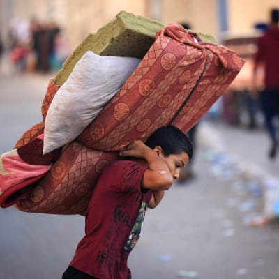 Poika kantaa patjaa ja petivaatteita selässään Gazassa.