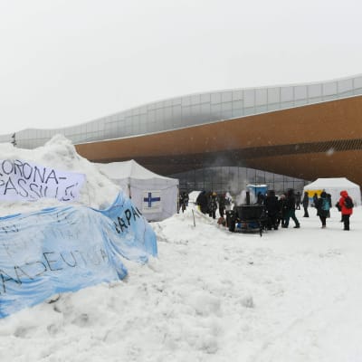 Personer står samlade vid tre vita tält på Medborgarplatsen. I förgrunden syns en snödriva där två banderoller ligger. Texten på banderollerna protesterar mot covidintyg.
