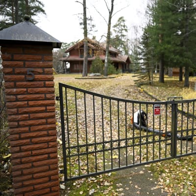 Ett hus i två plan syns på andra sidan en stängd port. Bakom grinden ligger en lövblåsare på vägen. Det är höst och det ligger löv på marken.