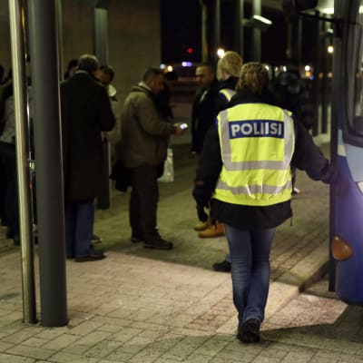 En polis med neongul väst står vid en buss och väntar på att en grupp asylsökande ska stiga på.