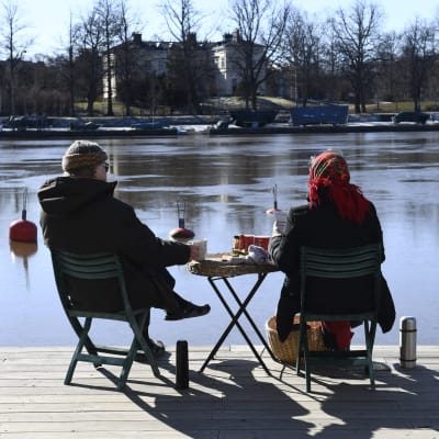 Två personer sitter på stolar med ett litet bord med mat emellan sig. Personerna blickar mot Kajsaniemi i Helsingfors.