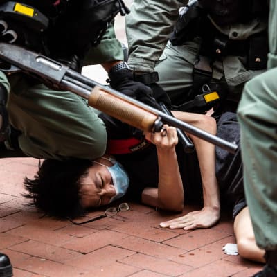 Poliser trycker ned en demonstrant mot marken. En av polisern ahåller ett gevär i handen.