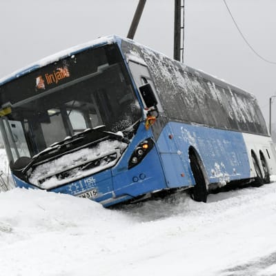 En buss har kört i ett dike på en snöig väg. Bussen är blå och vit och täckt av snö.