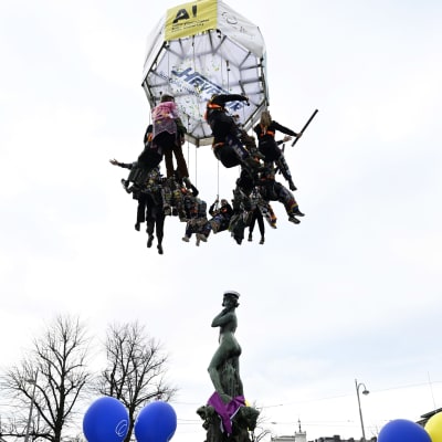 Studerande hänger i en ring ovanför Havis Amanda, som har fått sin mössa. Runt fontänen syns ett hav av människor med studentmössor och ballonger.