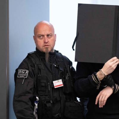 En väktare ledsagar en person som täcker sitt ansikte med en pärm och som är försedd med handklovar..