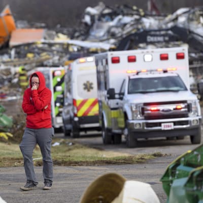 En person i röd luvtröja och grå byxor står framför en rad räddningsfordon. I bakgrunden syns delar av hus och bråte.
