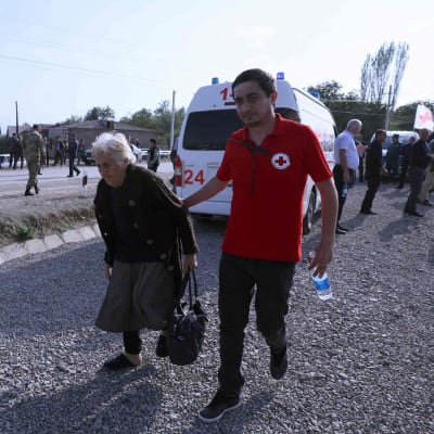 En man med ett rödakorsemblem på sin röda t-tröja leder en äldre kvinna längs en väg. Bakom dem står en ambulans och lite längre borta syns en vit flagga med ett rött kors.
