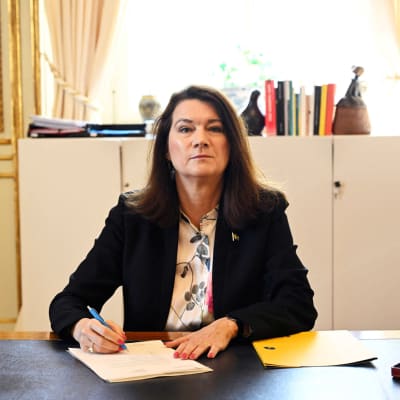 Ann Linde katsoo kameraan työpöydän takana. Kädessään hänellä on papereita ja kynä.