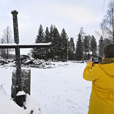 Keltaiseen talvitakkiin pukeutunut nainen kuvaa Rautjärven kirkon raunioita ja tulipalon jälkeen tapaninpäivänä portin pieleen nostettua kirkon ristiä 28. joulukuuta 2022. 